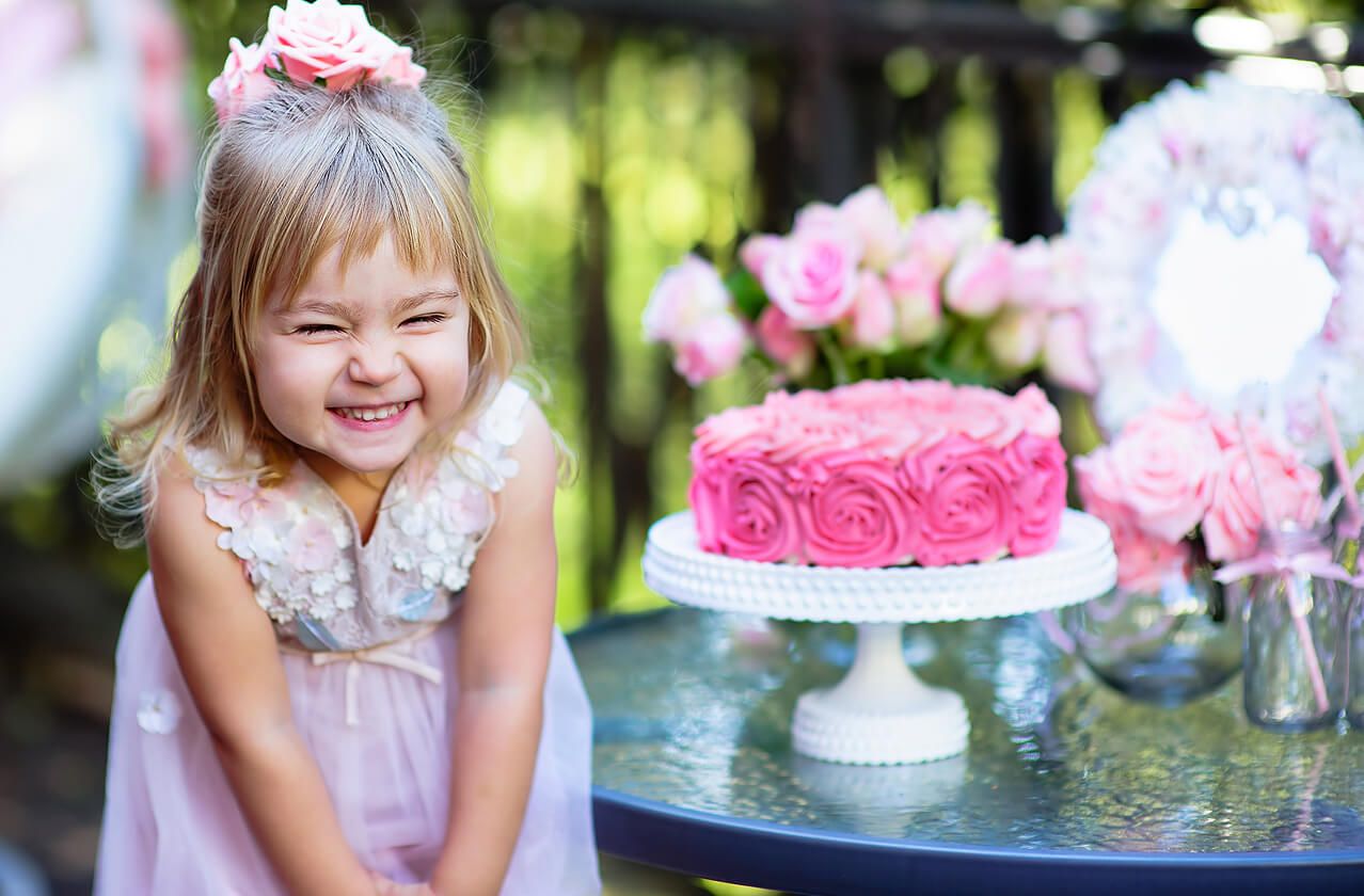 Ein kleines Mädchen in einem Kleid lächelt glücklich vor einem Kuchen und vielen Blumen.
